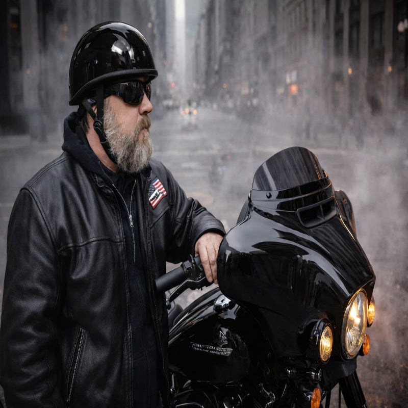 Man in black leather jacket and helmet standing next to a black motorcycle in an urban setting.